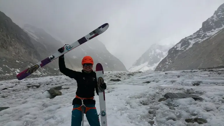 Een persoon met een oranje helm houdt een paar ski’s omhoog op een besneeuwde, rotsachtige berghelling, omringd door mist en hoge bergtoppen.